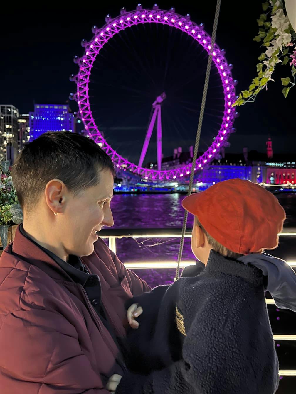 Carla and Baby O by the river Thames at night, a gleaming purple London Eye in the background between them